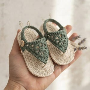 May include: A pair of small, handmade crochet sandals featuring a cream-colored sole and a sage green upper strap with a floral-inspired pattern. Each sandal is secured with a single light brown wooden button on the side and a heel strap.