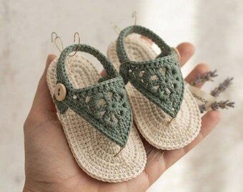 May include: A pair of small, handmade crochet sandals featuring a cream-colored sole and a sage green upper strap with a floral-inspired pattern. Each sandal is secured with a single light brown wooden button on the side and a heel strap.