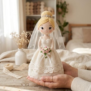 May include: A crocheted bride doll with blonde hair styled in a bun, wearing a white wedding dress and a sheer veil with small pearls. The doll holds a small bouquet of pink and white roses, featuring a silver-toned tiara on its head.