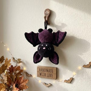 May include: A crocheted bat plush toy featuring a dark purple and black body with large wings, small round ears, and a tiny pink nose. The bat is suspended from a wooden wall hook above a rectangular wooden sign with the text HANDMADE WITH LOVE.