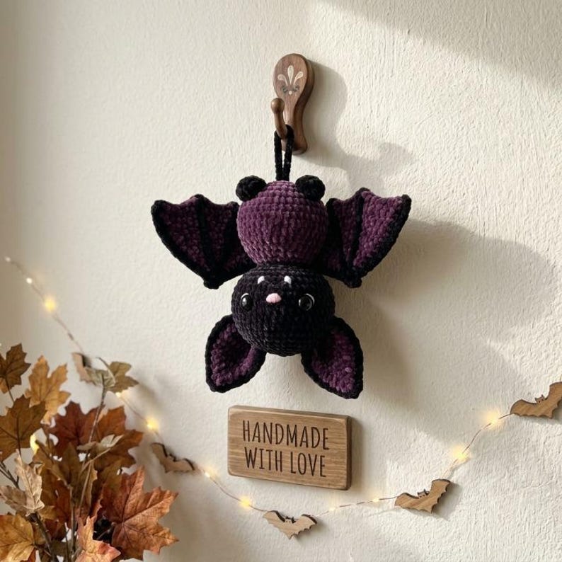 May include: A crocheted bat plush toy featuring a dark purple and black body with large wings, small round ears, and a tiny pink nose. The bat is suspended from a wooden wall hook above a rectangular wooden sign with the text HANDMADE WITH LOVE.