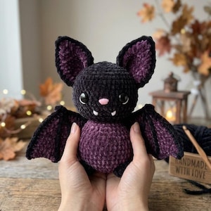 Könnte beinhalten: Ein gehäkeltes Fledermaus-Stofftier aus weichem schwarzem und dunkelviolettem Garn. Die Fledermaus hat große spitze Ohren mit violetten Innendetails, zwei schwarze Kunststoffaugen, eine kleine rosa Nase und zwei weiße Fangzähne im Mund.