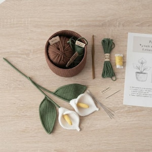 May include: A crochet kit featuring two white calla lilies with yellow spadices and green leaves, a brown yarn bowl containing brown and green yarn, a wooden crochet hook, a green yarn bundle, a small yellow yarn spool, a needle, and a paper pattern.