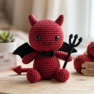 May include: A red crocheted devil doll with black wings, a black bowtie, and a black trident. The doll has two small horns, black eyes, and a stitched smile. A brown tag reads SPOOKY DEVIL CROCHET PATTERN, and a white card reads CRAFTY CORNER.