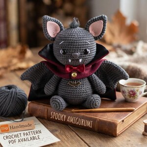 May include: A gray crocheted bat amigurumi with large ears, black eyes, and white fangs. It wears a dark red cape with a small bow tie and a gold bat-shaped brooch. The bat sits on a brown book titled Spooky Amigurumi next to a ball of gray yarn.