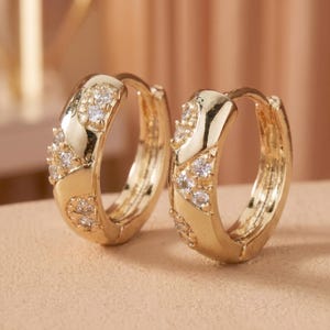 Op de afbeelding: Een paar gouden oorringen met een gepolijste metalen afwerking en ingebedde heldere, rondgeslepen stenen in een decoratief patroon langs de voorzijde van elke ring, voorzien van een scharnierend sluitingsmechanisme voor veilig dragen.