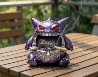 Puede incluir: Un cenicero morado con forma de Gengar y acabado metálico, con orejas puntiagudas, ojos rojos y una boca abierta que sirve de cuenco. El interior del cuenco está decorado con intrincados motivos plateados y contiene ceniza.