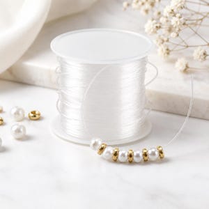 May include: A clear plastic spool of elastic cord featuring a strand of white round beads alternating with small metallic gold-toned spacer beads. The spool is cylindrical with a flat top and base, holding a continuous length of thin, transparent string.