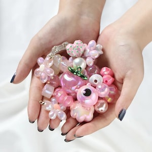 Op de afbeelding: Een verzameling diverse roze en iriserende kralen in verschillende vormen, waaronder bloemen, harten, bollen en een ronde karakterkraal met een opvallend zwart oog. De kralen hebben glanzende, doorschijnende en parelmoerachtige afwerkingen.