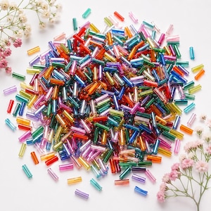 May include: A large collection of small, cylindrical glass tube beads in a vibrant rainbow of translucent colours, including red, orange, yellow, green, blue, and purple, scattered across a white surface with small sprigs of pink and white flowers.