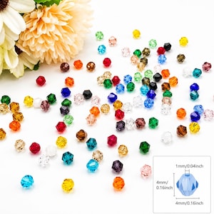 Pode incluir: Uma coleção de pequenas contas de cristal facetado multicoloridas espalhadas sobre uma superfície branca. Um diagrama no canto mostra uma conta azul com dimensões de 1 mm por 4 mm. Uma grande flor amarelo-pálida é visível.