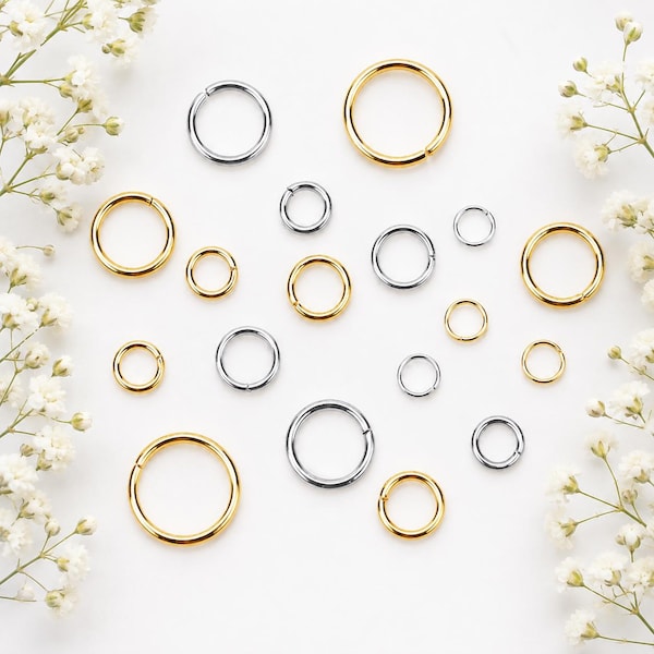 Puede incluir: Una colección de anillas de salto de metal en tonos dorados y plateados, con diversos diámetros y grosores. Estos fornituras circulares para joyería están dispuestas en un patrón disperso, mostrando superficies lisas y pulidas.