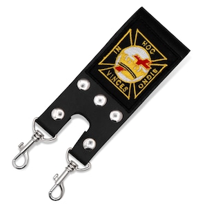 Op de afbeelding: Een zwarte leren riem met vijf zilverkleurige metalen klinknagels en twee zilverkleurige draaibare karabijnhaken aan de onderkant. Een geborduurde patch is in het midden geplaatst met een gouden kruis en de tekst IN HOC SIGNO VINCES.
