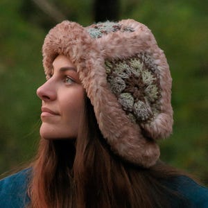 May include: A trapper-style hat featuring a plush, dusty pink faux fur trim and a crown constructed from crocheted granny squares in muted shades of sage green, brown, and cream. The hat has ear flaps and a soft, textured appearance.