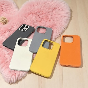 Könnte beinhalten: Eine Sammlung von fünf Smartphone-Hüllen in matten Unifarben, darunter Schwarz, Grau, Weiß, Gelb und Orange. Jede Hülle verfügt über einen rechteckigen Kameraausschnitt an der oberen linken Ecke sowie präzise Öffnungen für Anschlüsse.
