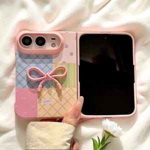 Op de afbeelding: Een opvouwbaar smartphonehoesje in pastelroze, blauw en geel met een patchworkpatroon. De achterkant heeft een verhoogde roze strik, een camera-uitsparing en het woord LUCKY in kleine letters. Het hoesje heeft een matte afwerking.