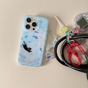 May include: A light blue smartphone case featuring a pattern of small fish and a black cat silhouette. Attached to the side is a beaded charm with a miniature clear bag containing a blue fish, a yellow lollipop, and a digital pet device.