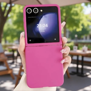 May include: A bright pink foldable smartphone case featuring a cutout for the dual camera lenses and a secondary display screen. The screen shows the time 21:24, the date, and various status icons. The case has a smooth, matte finish.
