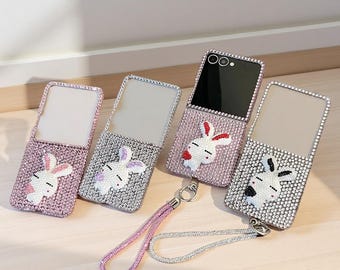 May include: Four rectangular phone cases with mirrors, each covered in small sparkling crystals. Three cases feature a white rabbit design with colored accents, while one case is attached to a phone and includes a matching crystal wrist strap.