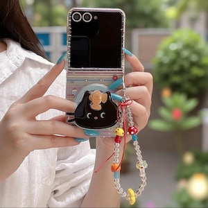 May include: A foldable smartphone case featuring a striped pastel background with small star and heart icons. A black cat-shaped grip is attached to the centre, topped with a small fish. A beaded phone charm with mushroom and fruit accents hangs from the side.