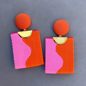 Op de afbeelding: Een paar oorbellen met een ronde, oranje, met stof beklede knop verbonden aan een rechthoekige hanger. De hanger heeft een getextureerd oppervlak met een golvend patroon in felroze en oranje, geaccentueerd door een goudkleurig halfrond stuk.