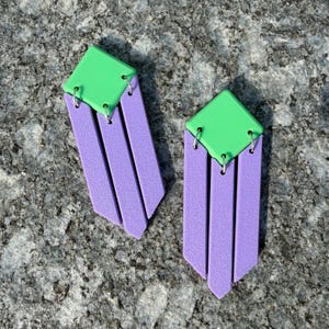 Op de afbeelding: Een paar hangende oorbellen met een felgroen ruitvormig bovendeel verbonden aan drie verticale, rechthoekige paarse staven. De onderdelen zijn met elkaar verbonden door kleine zilverkleurige metalen ringetjes voor een geometrisch ontwerp.