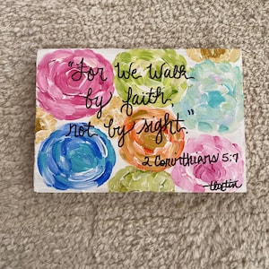 May include: A colorful painted wooden sign with the quote "For we walk by faith, not by sight" from 2 Corinthians 5:7. The sign has a floral design with pink, blue, green, and orange flowers.