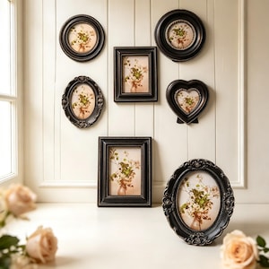 May include: A collection of six black picture frames in various shapes including oval, rectangular, and heart-shaped, each containing a print of a floral arrangement. The frames feature ornate, textured borders and are displayed on a wall.