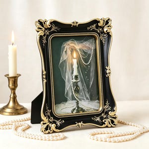 May include: A rectangular picture frame with a matte black finish and ornate gold-coloured scrollwork detailing. The frame contains an image of a lit candle, a sheer white veil, and a string of white pearls against a dark green background.