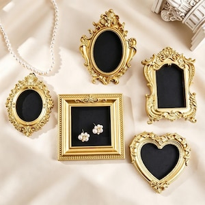 May include: A collection of five ornate gold-colored frames with black interiors, including oval, rectangular, and heart-shaped designs. One square frame contains a pair of white pearl stud earrings. The frames feature intricate scrollwork details.