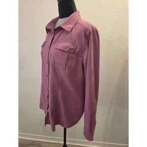 Puede incluir: Una camisa de manga larga abotonada en color rosa polvoriento con cuello clásico, dos bolsillos en el pecho con solapas y tapeta frontal con botones. La camisa incluye mangas largas con pestañas para enrollar y un dobladillo curvo.