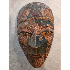 May include: A hand-carved wooden mask featuring intricate batik-style patterns in shades of dark green, brown, and tan. The mask displays almond-shaped eyes, a prominent nose, and a pointed chin, with detailed floral and geometric motifs.