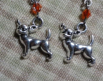 May include: Silver dangle earrings featuring a small silver chihuahua charm and an orange crystal bead.