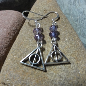 May include: A pair of silver-toned earrings featuring the Deathly Hallows symbol. Each earring has a triangular charm with a circle and vertical line, and is accented with purple beads. The earrings hang from a simple hook.