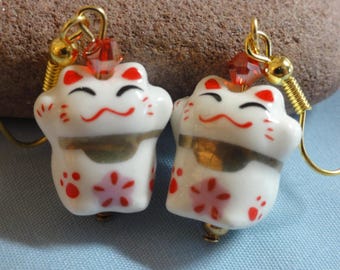 May include: A pair of white ceramic cat earrings with red and gold accents. The cats are depicted with raised paws and a smiling face. The earrings are suspended from gold hooks with red crystal beads.
