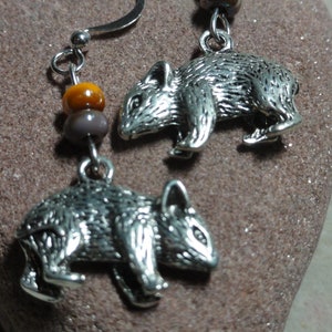 May include: Silver dangle earrings featuring a small, detailed animal charm. The charm resembles a wombat. The earrings have a brown and orange bead on the top.