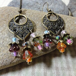 May include: A pair of antique brass earrings with a filigree design. Each earring has a decorative pendant with dangling colourful glass beads in shades of purple, orange, green, and pink. The earrings are displayed on a textured stone surface.