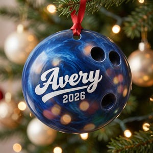 May include: A spherical Christmas ornament shaped like a blue marbled bowling ball with three finger holes. The name Avery is printed in white script above the year 2026 in white block text. A thin red ribbon is attached to the top for hanging.