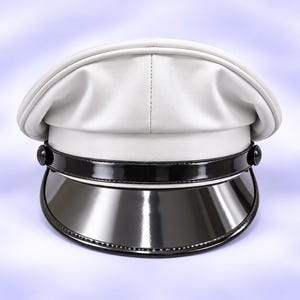 Pode incluir: Um boné de estilo militar branco com uma coroa branca lisa e mate, contrastada por uma pala de verniz preto. Uma tira de queixo em verniz preto está fixada na parte frontal com dois pequenos botões pretos redondos em cada lado.