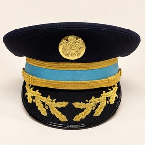 Può includere: Un berretto da servizio militare blu scuro con un emblema centrale a forma di aquila dorata, una fascia azzurra e un sottogola intrecciato dorato. La visiera nera è decorata con foglie di quercia ricamate in oro su entrambi i lati.