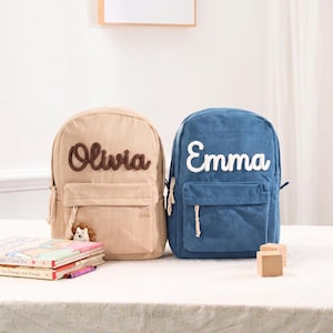 May include: Two corduroy backpacks, one beige and one blue, each featuring a front pocket and a name embroidered in cursive script. The beige backpack displays the name Olivia in brown, while the blue backpack displays the name Emma in white.