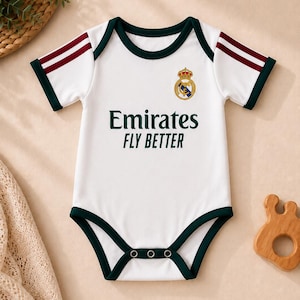May include: A white short-sleeve baby bodysuit featuring dark green trim on the neckline, sleeves, and leg openings. The chest displays the Real Madrid crest and the text Emirates FLY BETTER in dark green. Two maroon stripes accent each sleeve.