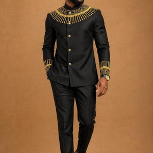 K&ouml;nnte beinhalten: Ein schwarzer Anzug bestehend aus einer Jacke mit Stehkragen, die am Ausschnitt und an den &Auml;rmelb&uuml;ndchen mit goldener Stickerei verziert ist, kombiniert mit einer passenden schwarzen Hose. Die Jacke hat eine Knopfleiste mit goldenen Kn&ouml;pfen.