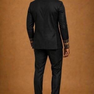 K&ouml;nnte beinhalten: Ein schwarzer Anzug bestehend aus einem taillierten Blazer und einer passenden Hose. Der Blazer hat lange &Auml;rmel mit aufwendigen goldenen Stickereien an den Manschetten. Das Ensemble wird mit schwarzen Slippern aus Glattleder kombiniert.
