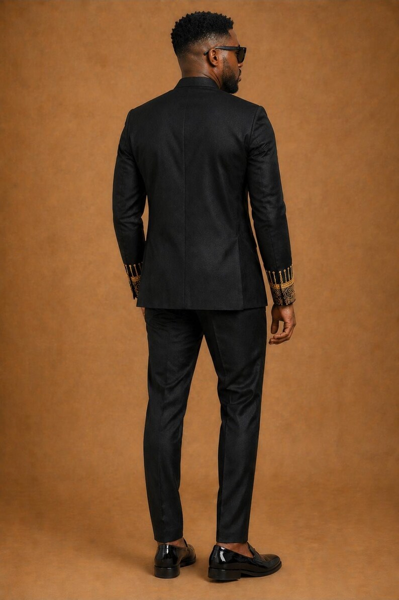 K&ouml;nnte beinhalten: Ein schwarzer Anzug bestehend aus einem taillierten Blazer und einer passenden Hose. Der Blazer hat lange &Auml;rmel mit aufwendigen goldenen Stickereien an den Manschetten. Das Ensemble wird mit schwarzen Slippern aus Glattleder kombiniert.
