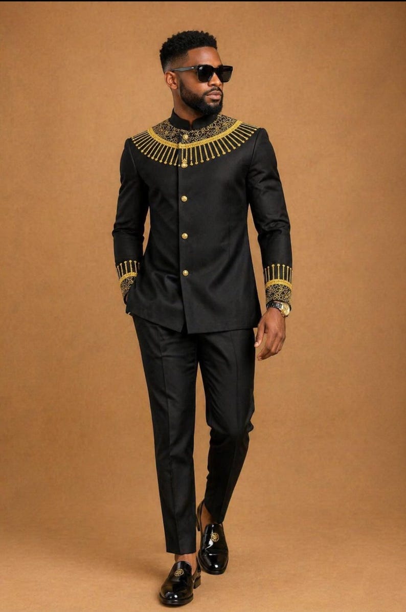 K&ouml;nnte beinhalten: Ein schwarzer Anzug bestehend aus einer Jacke mit Stehkragen, die am Ausschnitt und an den &Auml;rmelb&uuml;ndchen mit goldener Stickerei verziert ist, kombiniert mit einer passenden schwarzen Hose. Die Jacke hat eine Knopfleiste mit goldenen Kn&ouml;pfen.