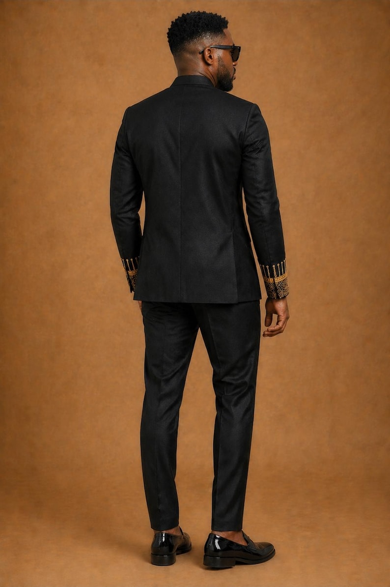 K&ouml;nnte beinhalten: Ein schwarzer Anzug bestehend aus einem taillierten Blazer und einer passenden Hose. Der Blazer hat lange &Auml;rmel mit aufwendigen goldenen Stickereien an den Manschetten. Das Ensemble wird mit schwarzen Slippern aus Glattleder kombiniert.