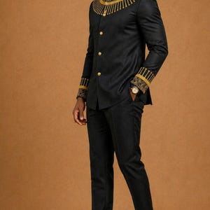 K&ouml;nnte beinhalten: Ein schwarzes Set aus lang&auml;rmeliger Jacke und passender Hose. Die Jacke hat einen Stehkragen mit goldener Stickerei, goldene Kn&ouml;pfe und gold gemusterte Manschetten. Das Ensemble wird mit schwarzen Slippern und einer Sonnenbrille kombiniert.