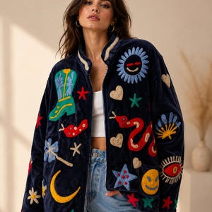May include: A navy blue zip-up jacket featuring colorful embroidered patches including a cowboy boot, a sun, a snake, a crescent moon, hearts, stars, a smiley face, and an eye. The jacket has a high collar and is worn over a white top and blue jeans.
