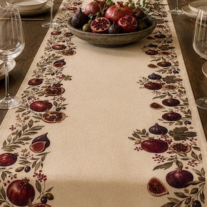 May include: A cream-colored table runner featuring a botanical print of pomegranates, figs, and green leaves along both edges. The design includes whole and sliced fruits in deep red and purple tones, arranged in a repeating pattern on fabric.